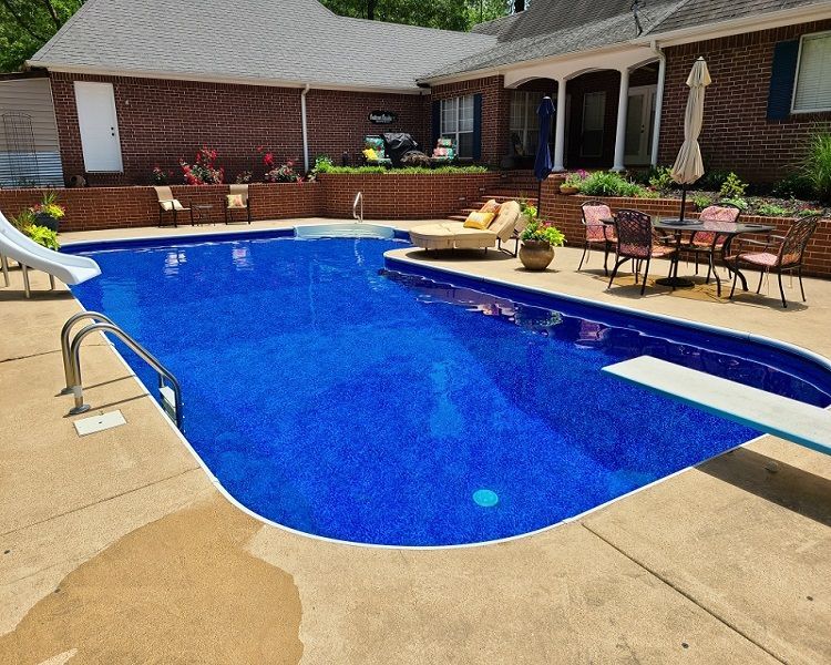 Blue-tiled swimming pool surrounded by a concrete patio. House with brick exterior is visible in the background.