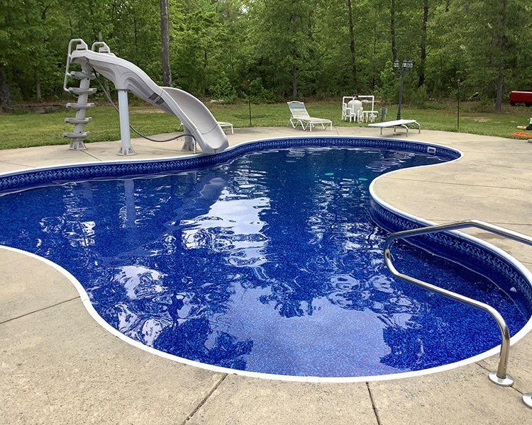 A blue-tiled, kidney-shaped swimming pool with a slide, surrounded by concrete, lawn, and trees.