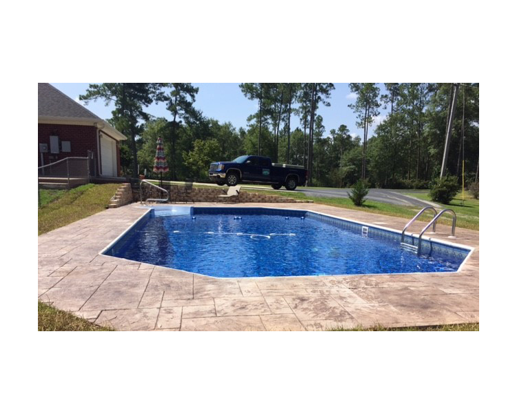 Rectangular blue tiled pool with concrete border, set in a grassy yard near a road and trees.