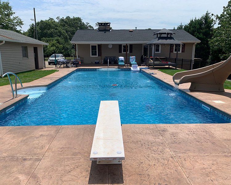 Rectangular pool with diving board and water slide in a backyard. House and small building in the background. Sunny day.