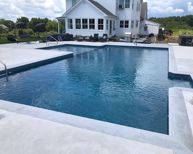 Blue swimming pool with concrete deck next to a two-story white house on a sunny day.