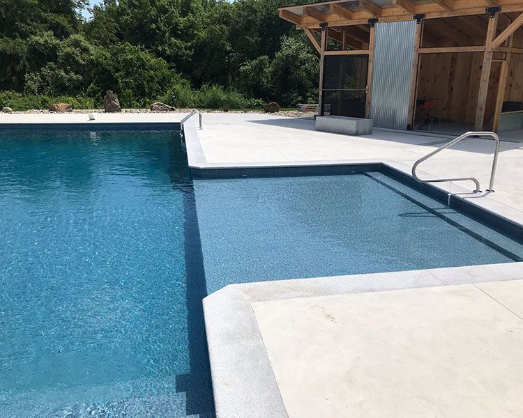 A swimming pool with blue water and concrete decking. A wooden structure is in the background.