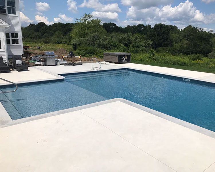 Pool with light concrete decking, blue water, and view of a treeline under a partly cloudy sky.