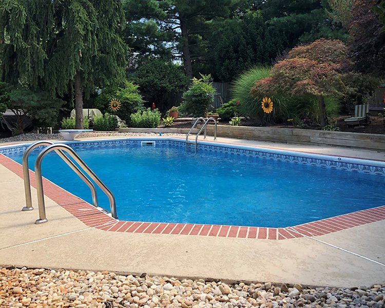 A rectangular swimming pool with blue water and brick coping surrounded by a concrete patio and landscaping.