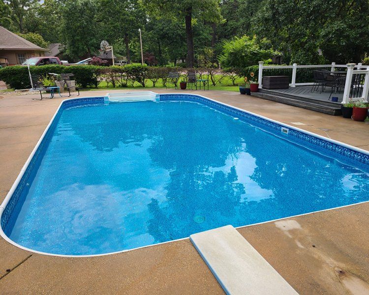 Rectangular swimming pool with blue water and diving board, concrete deck, and wooden deck with furniture.