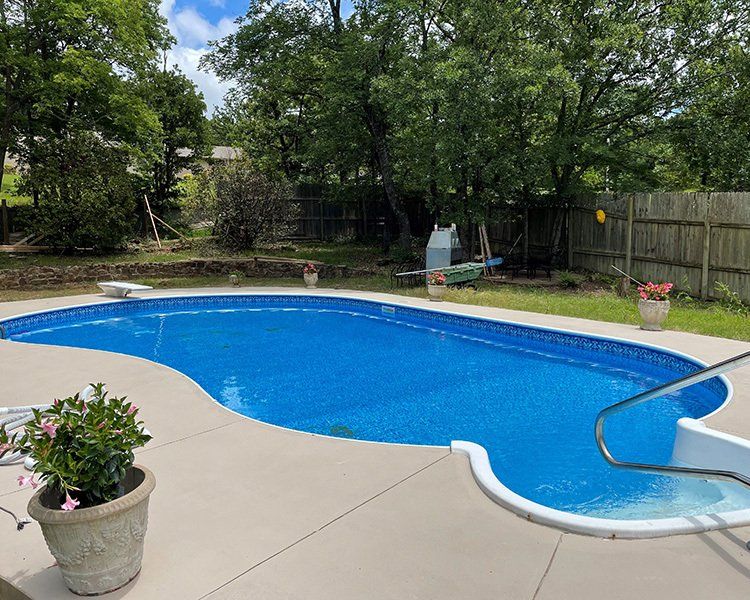 An in-ground, blue-tiled swimming pool surrounded by concrete, with a green lawn and trees in the background.