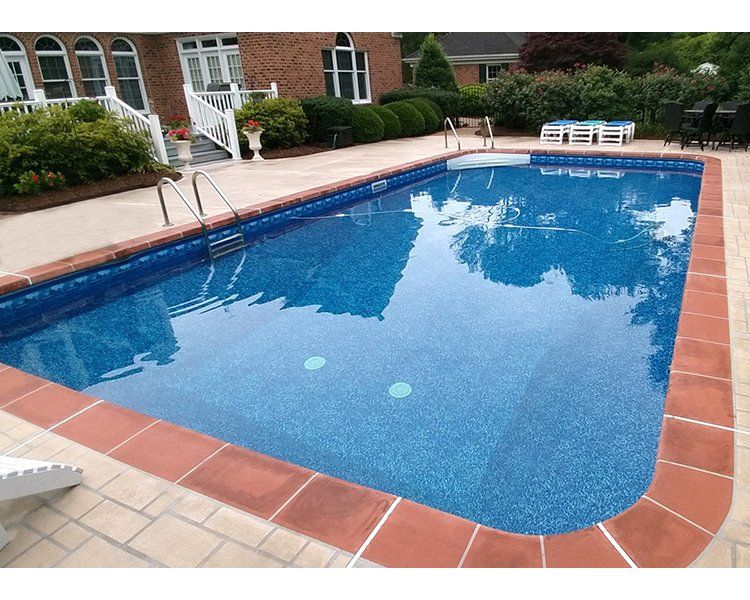 Rectangular blue pool with red brick border, concrete patio, near a house with landscaping.