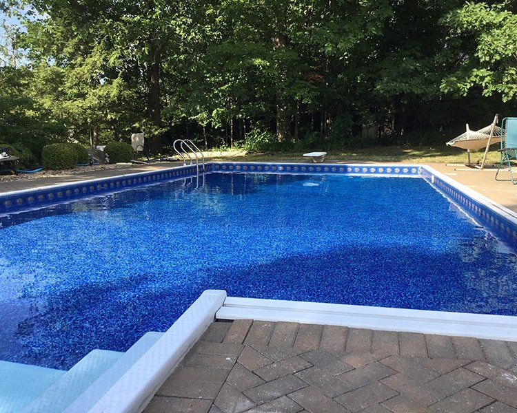 Swimming pool with blue mosaic-tiled interior, surrounded by a brick patio. Trees in background.