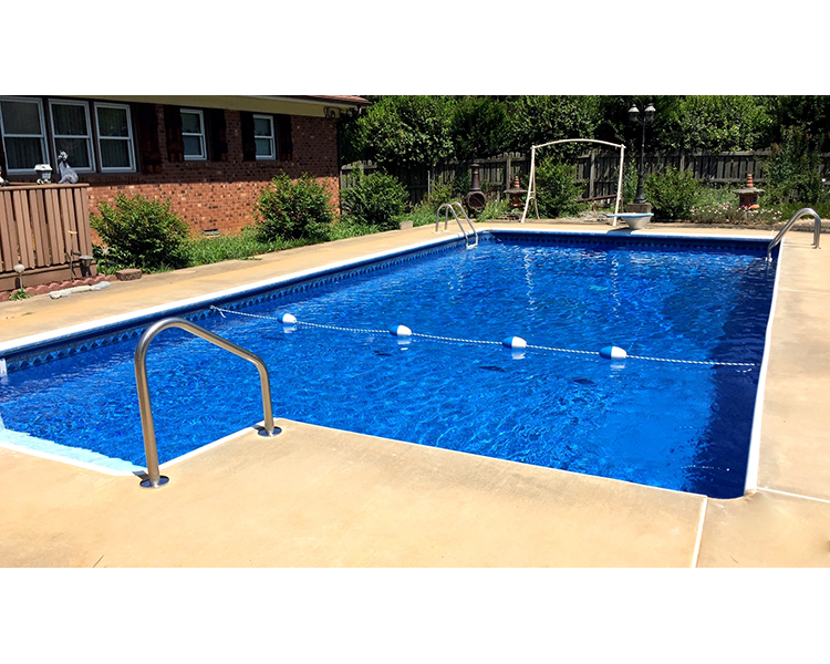 Swimming pool, blue water, tan concrete deck, stainless steel ladder, brick house in the background.