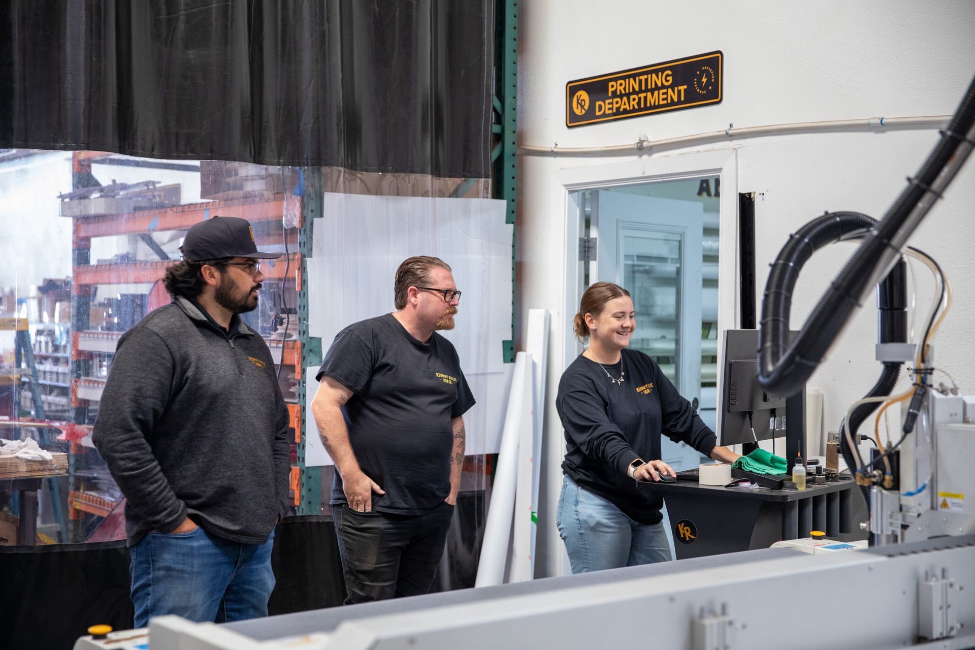Three people observing a CNC machine in a workshop, with a woman operating the controls.