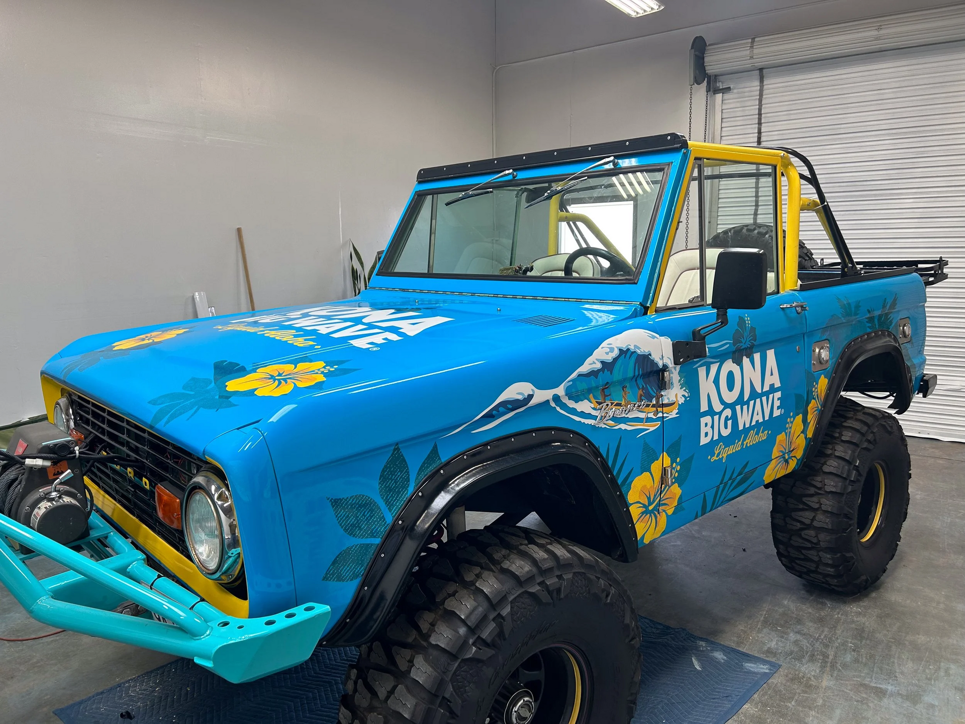 Blue and yellow vintage Bronco truck with Kona Brewing graphics, large tires, and roll cage.