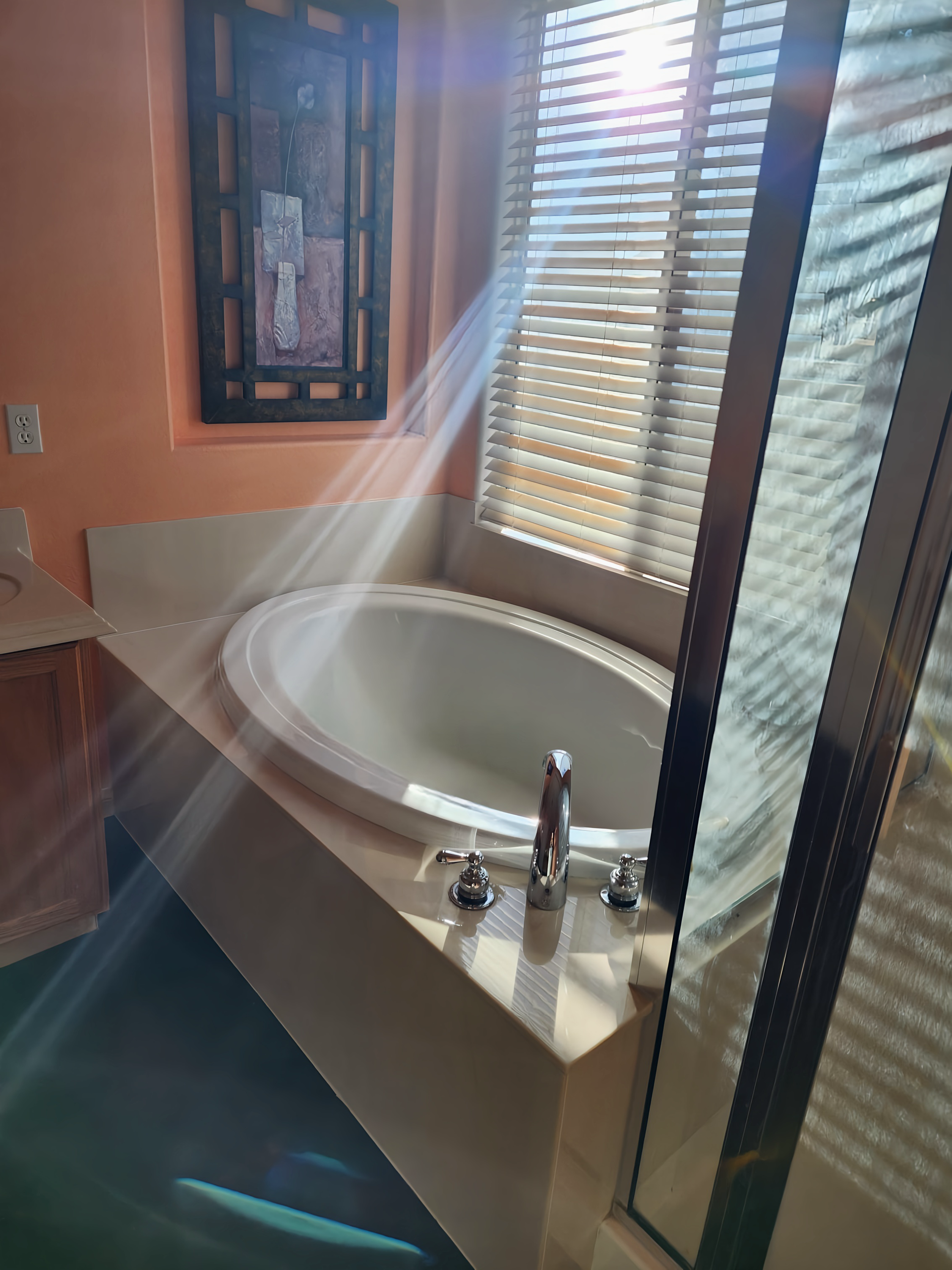 Bathroom with oval tub, beige countertop, blinds, art, and sunlight streaming through.