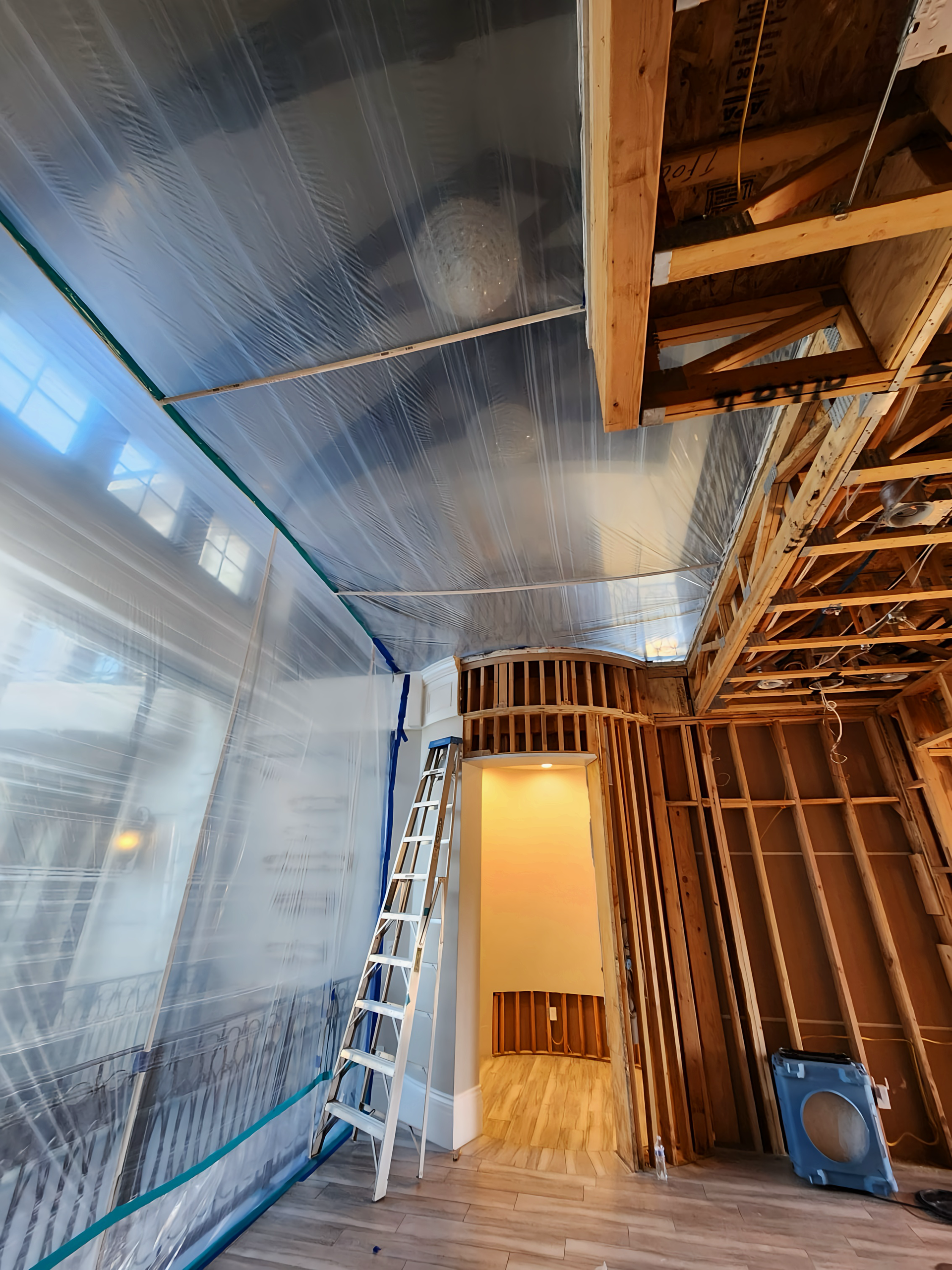 Interior during renovation: plastic sheeting covers ceiling, framing exposed, ladder, open doorway, bright light.