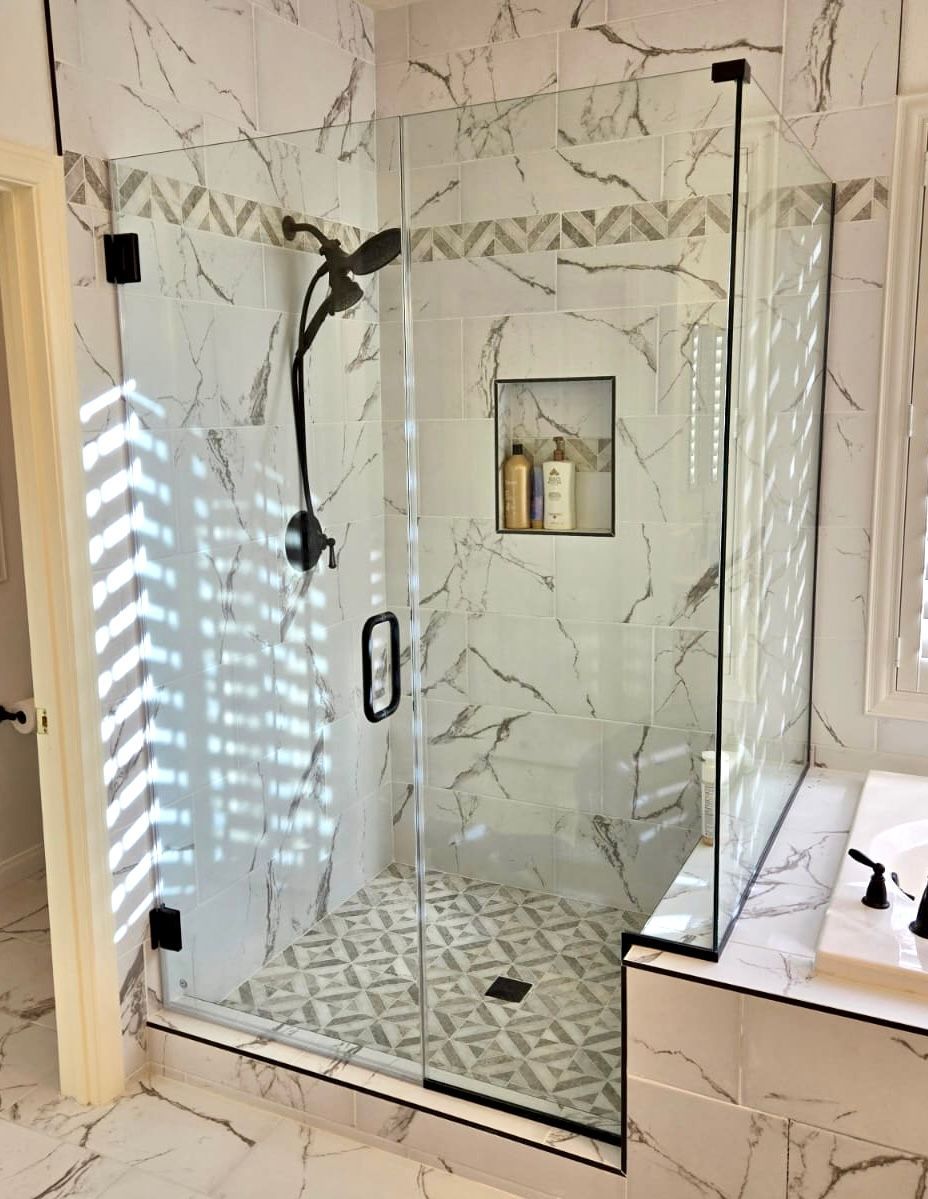Modern marble-tiled shower with a glass door. Black hardware and a built-in shelf.