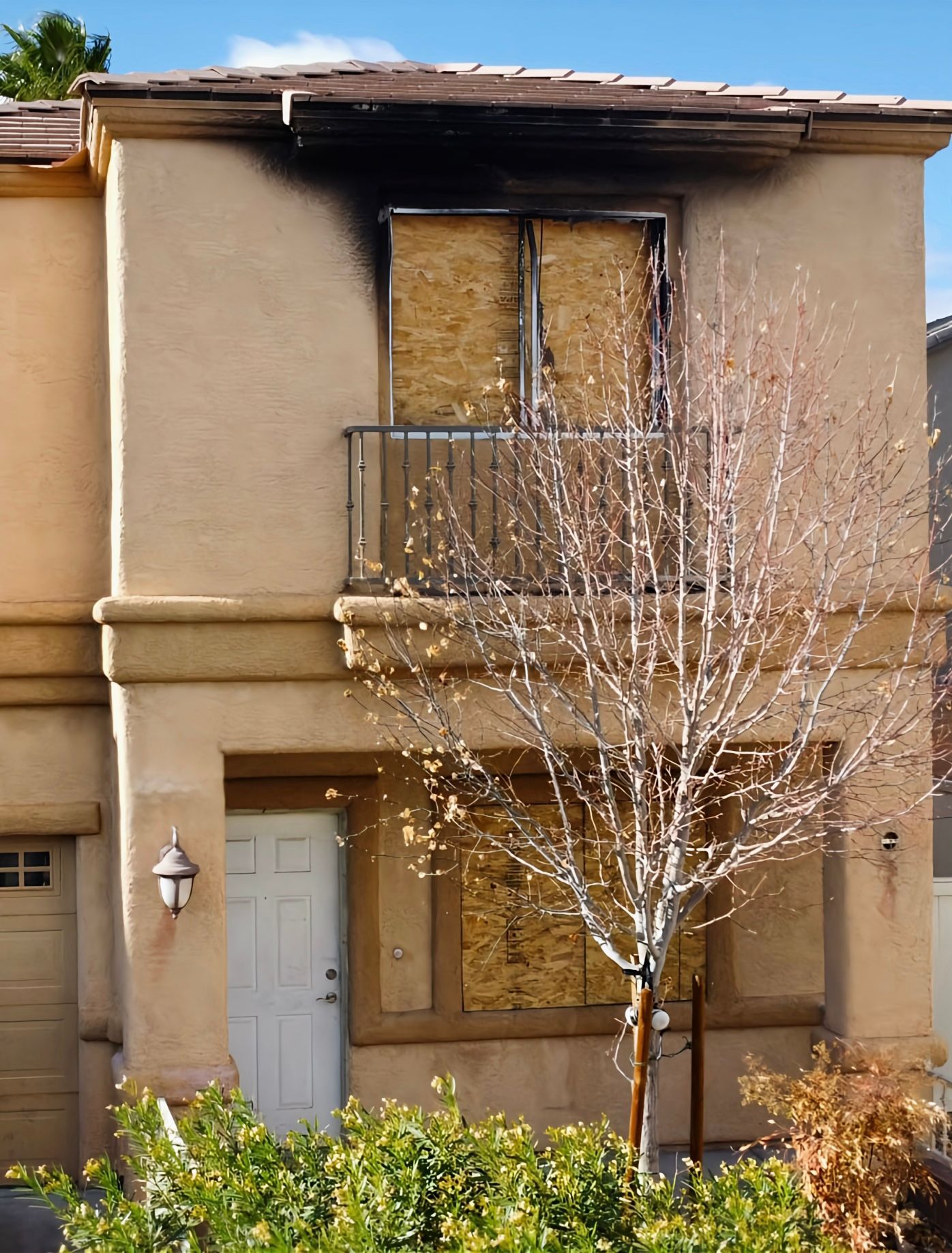 Two-story house with boarded-up windows and fire damage above. A bare tree stands in front, and the sky is blue.