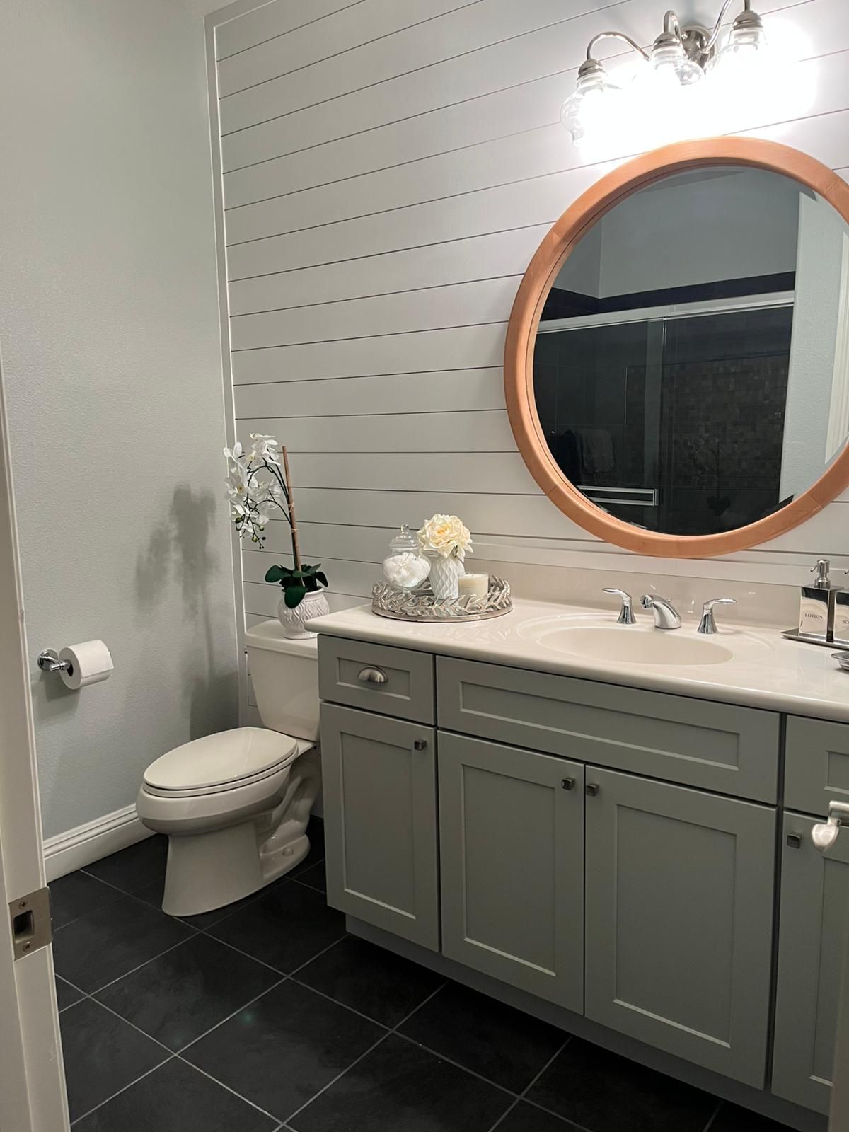 A bathroom featuring a gray vanity, white countertop, round mirror, and a shiplap accent wall.