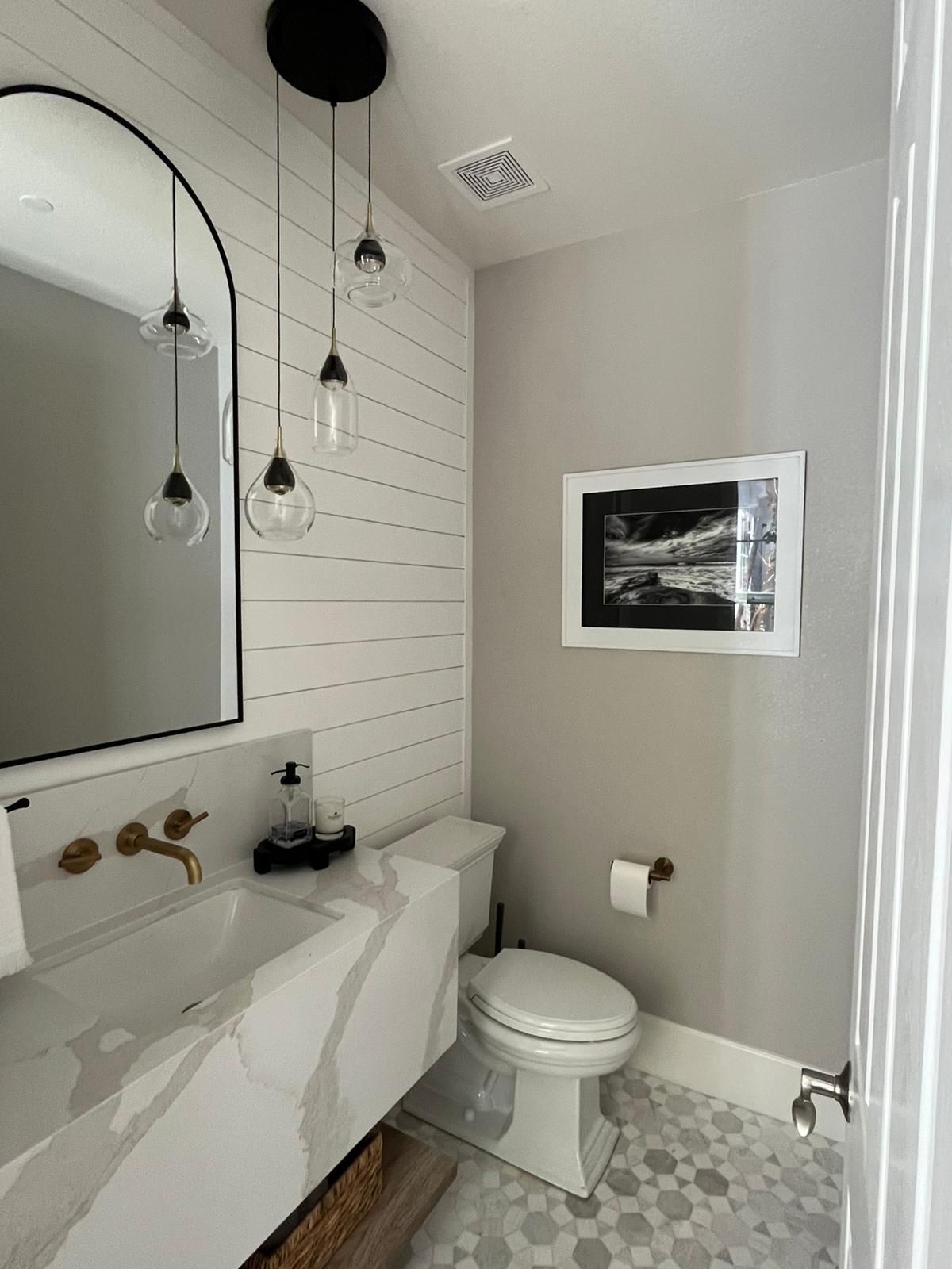 A modern bathroom with a white marble countertop, a shiplap wall, and an arched mirror.