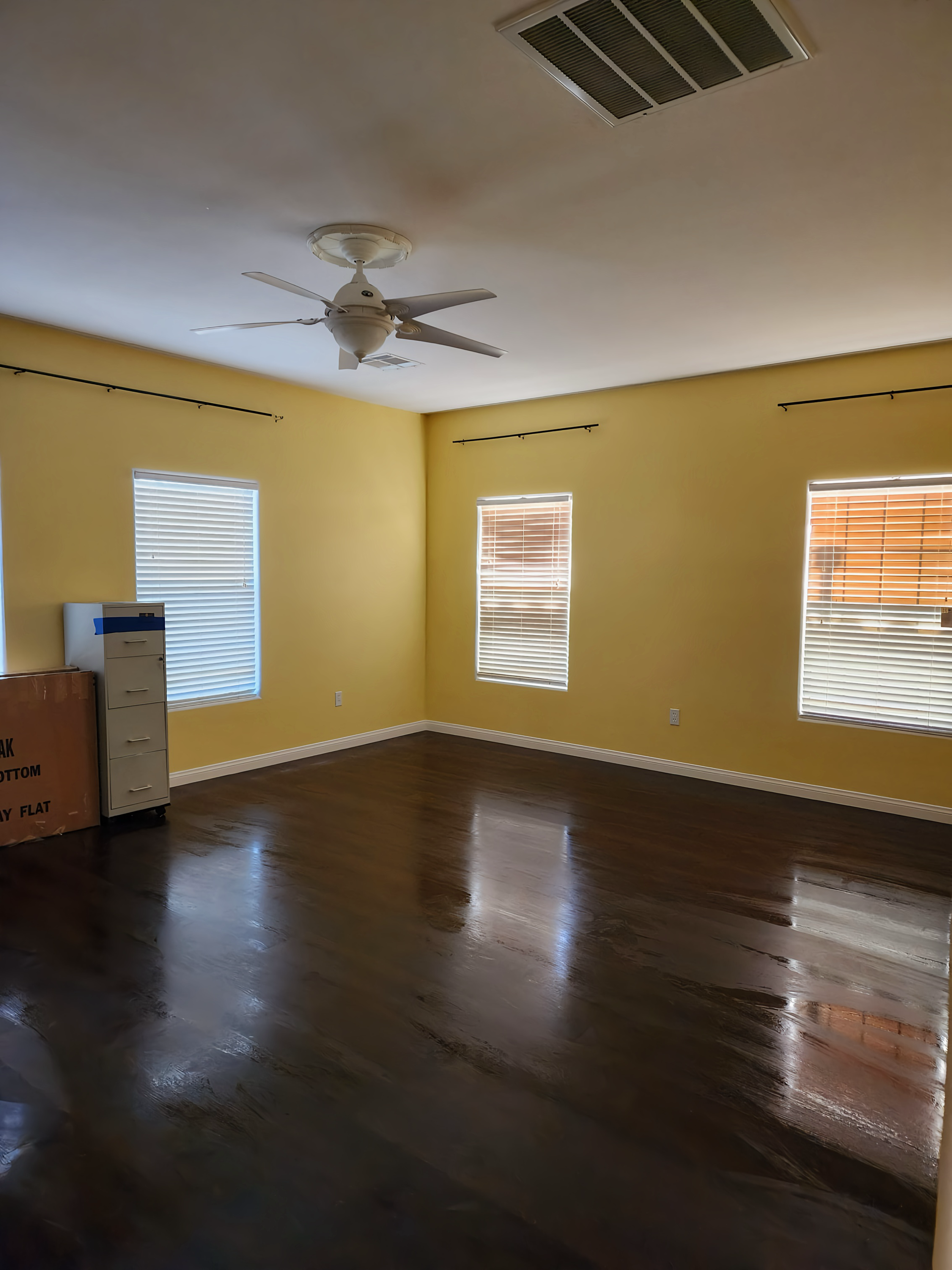 Empty room with dark hardwood floors, yellow walls, and white trim; windows with blinds.