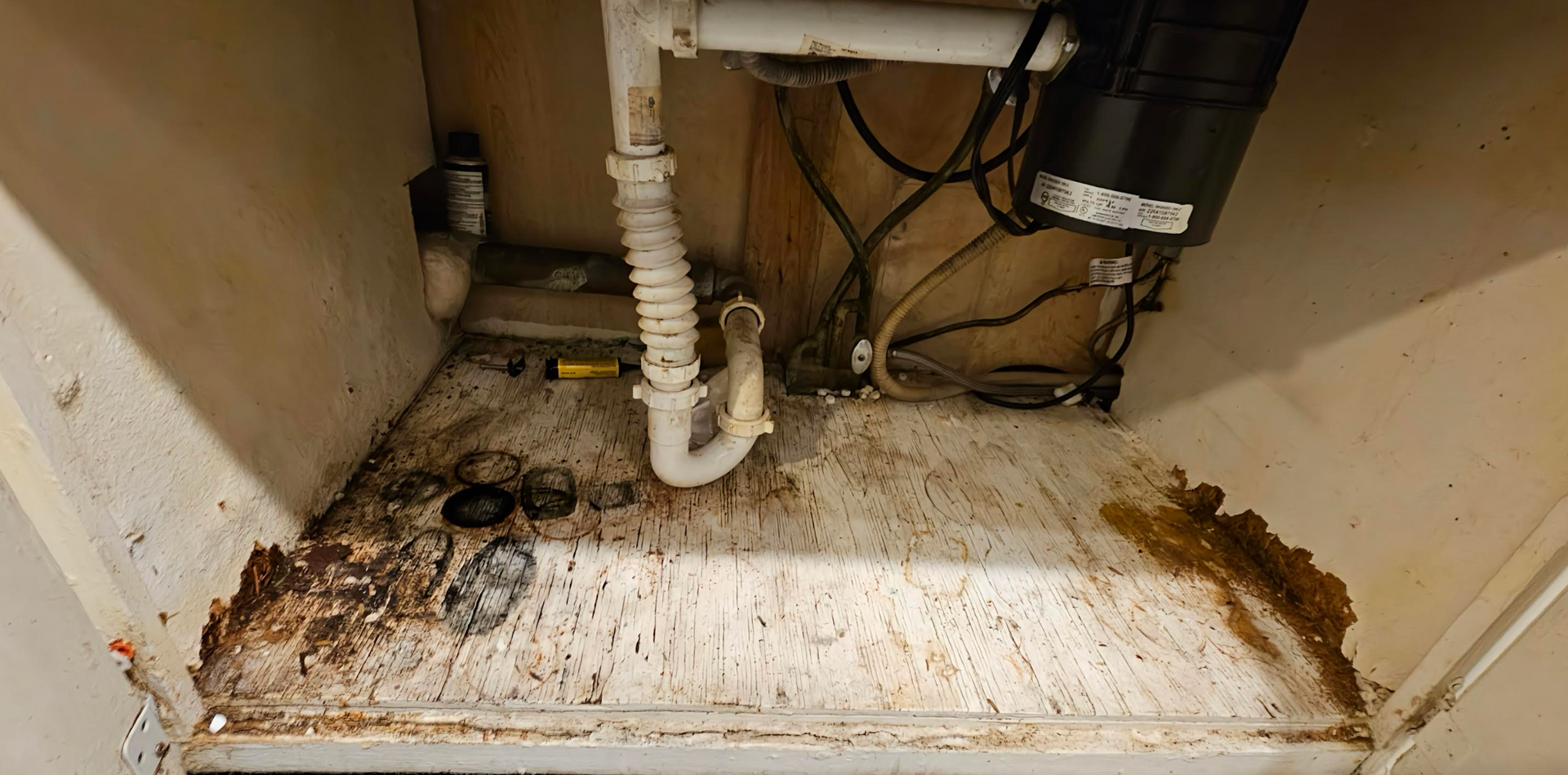 Under sink cabinet interior, with plumbing, garbage disposal, and debris-covered surface; noticeable staining and damage.