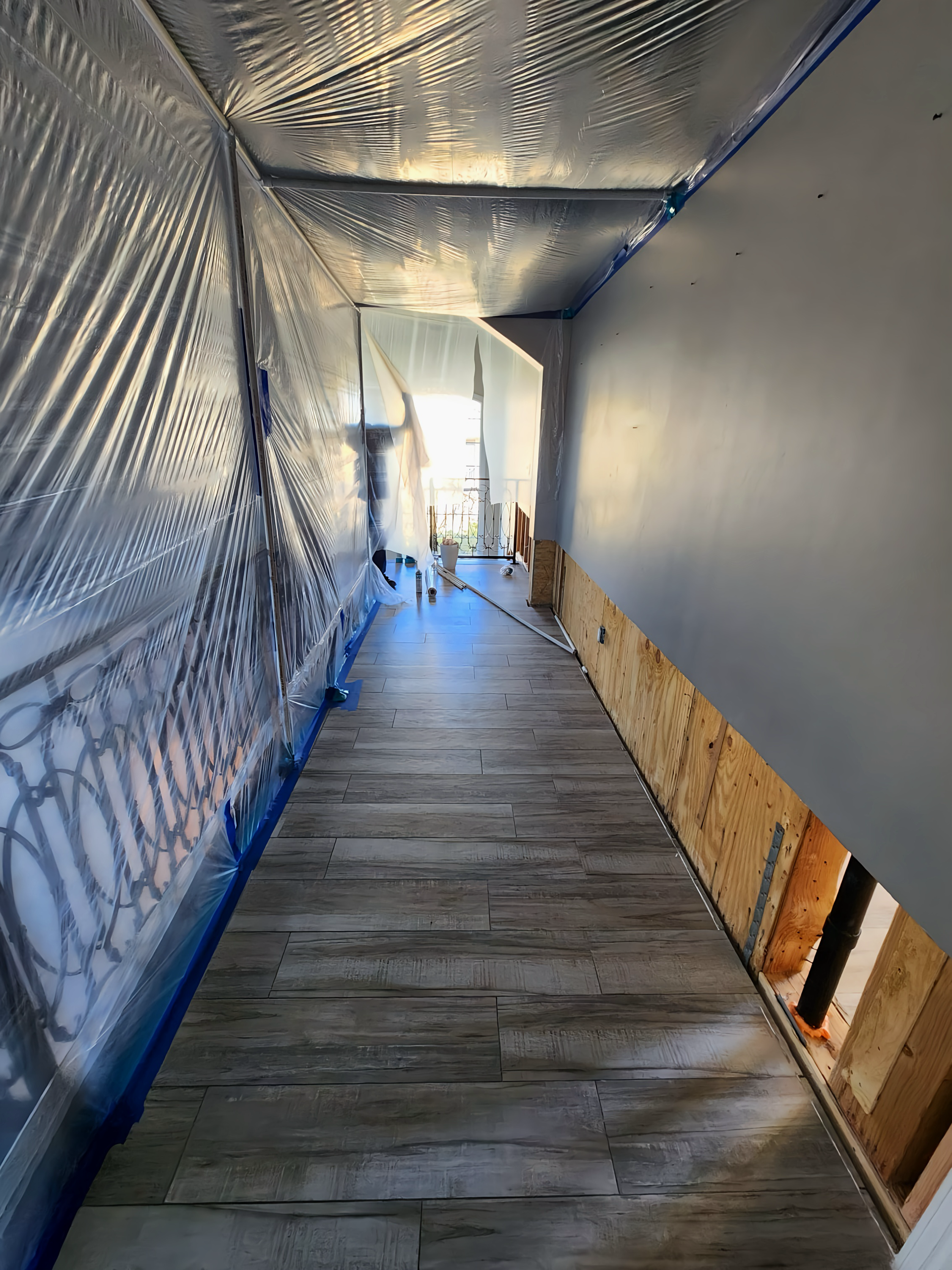 Narrow hallway under construction; wood-look tile floor, plastic sheeting, unfinished walls.
