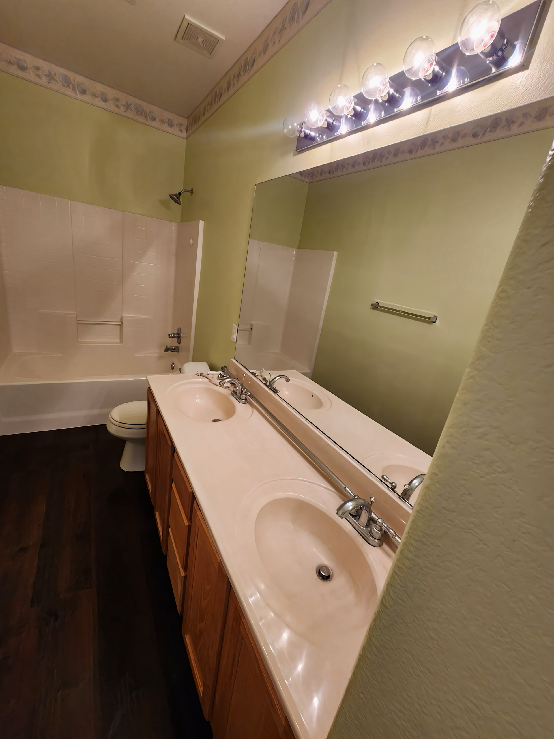 Bathroom with double sinks, large mirror, and tub. Light green walls, dark wood floor, and tan countertop.