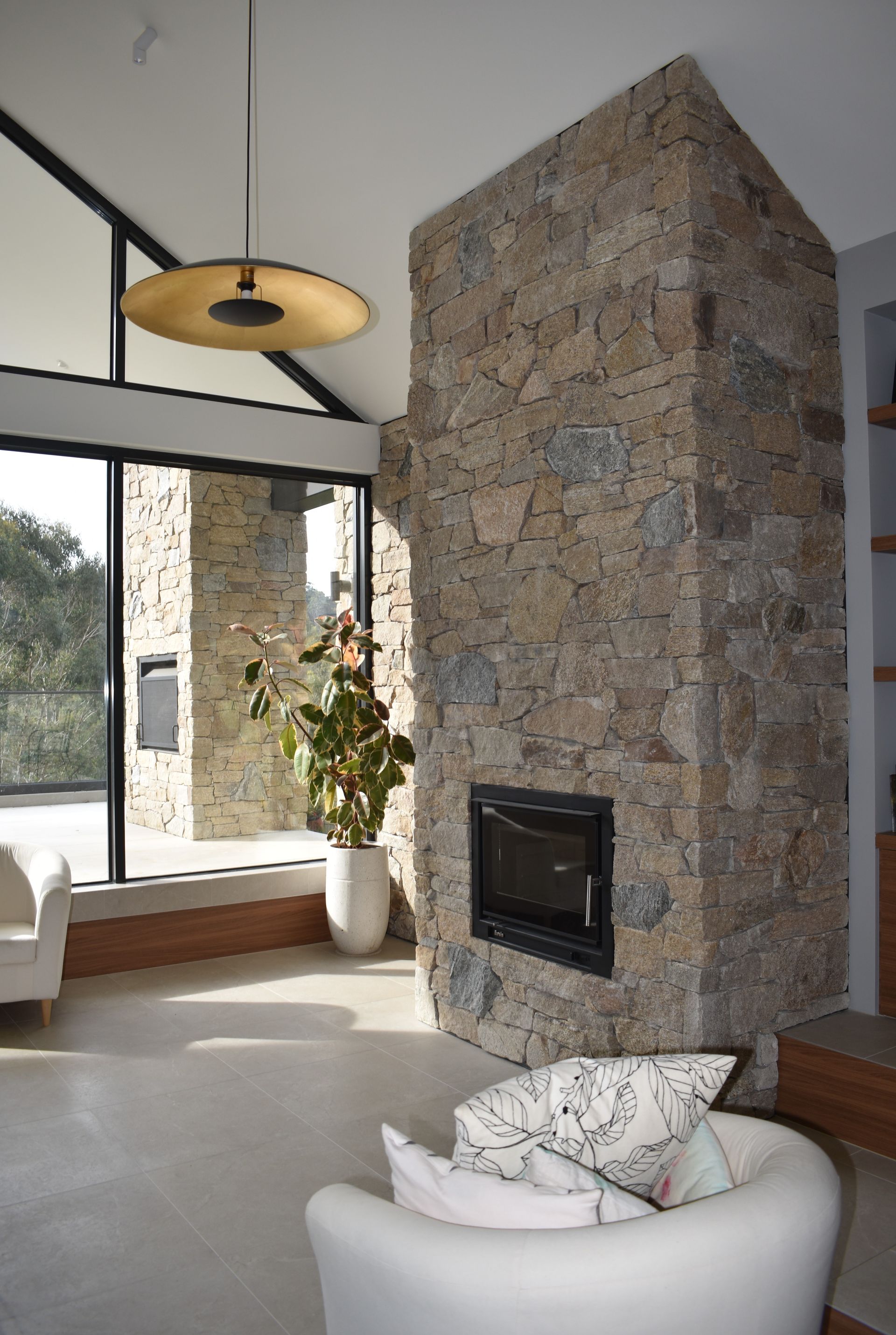 Modern living room with stone fireplace, large windows, and a white lounge chair with pillows