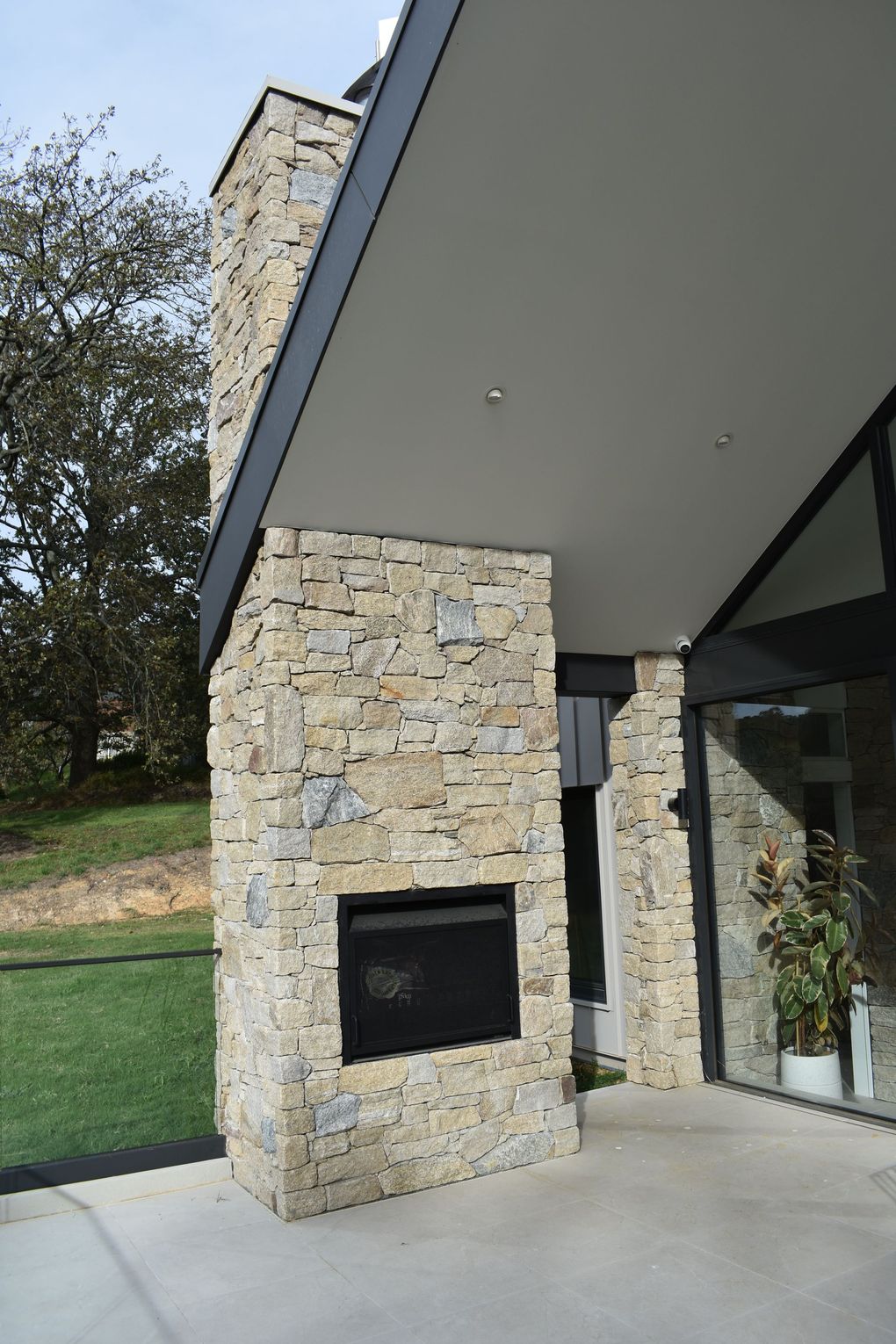 Modern stone fireplace under a slanted roof beside glass doors, with a concrete patio and grassy slope nearby