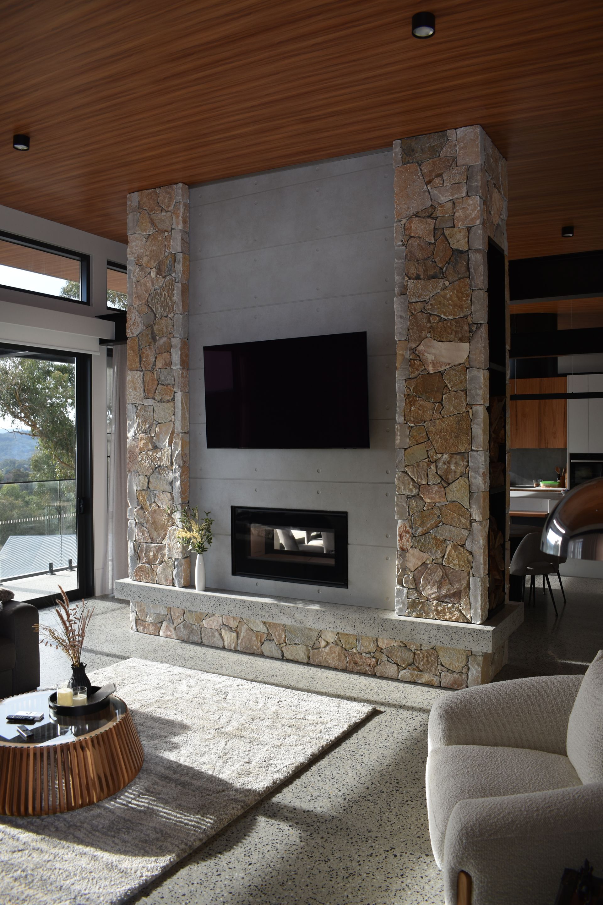 Modern living room with stone fireplace, wall-mounted TV, and large windows with sunlight.