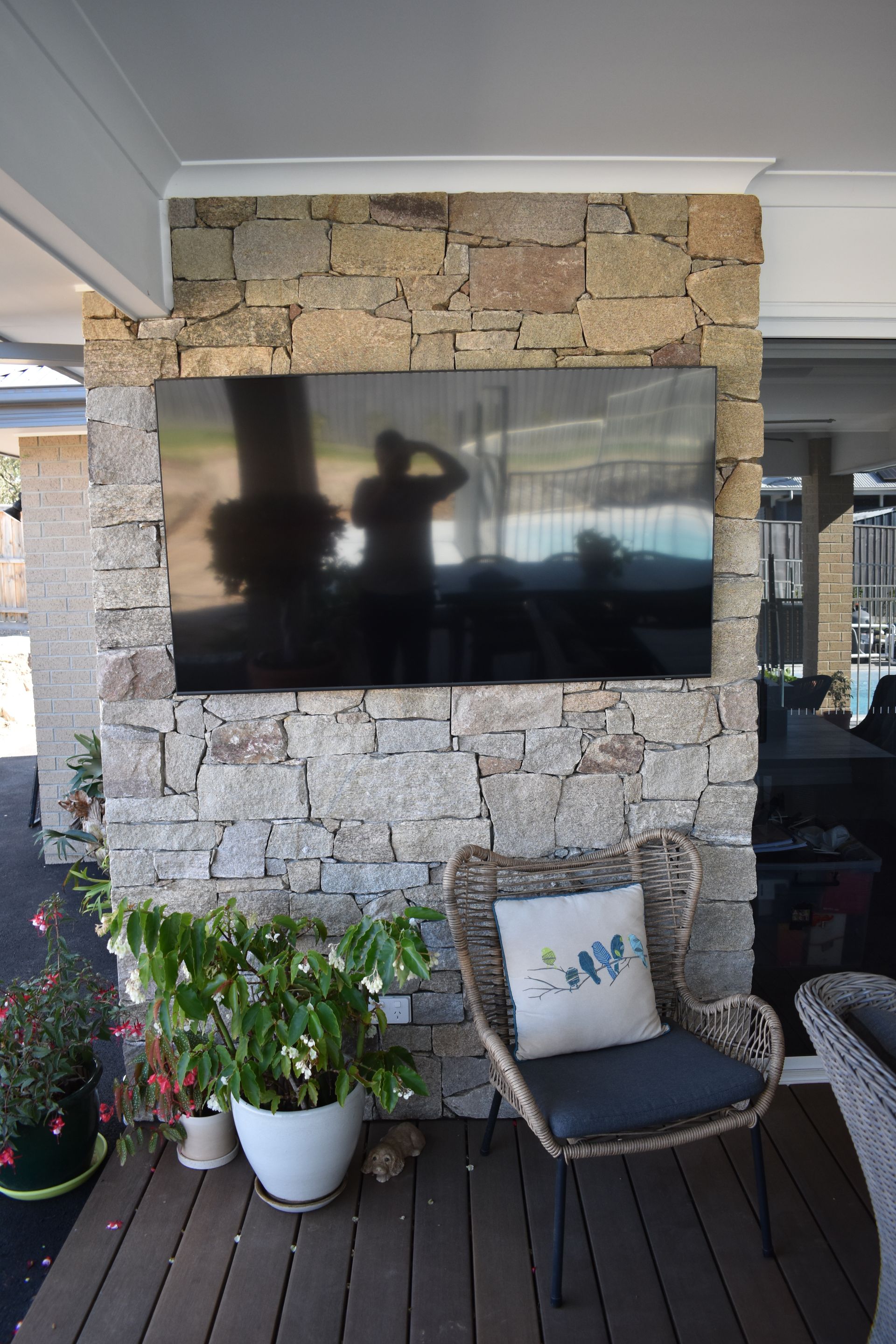 Outdoor patio with stone wall-mounted TV, potted flowers, and a black chair with pillow on a wooden deck