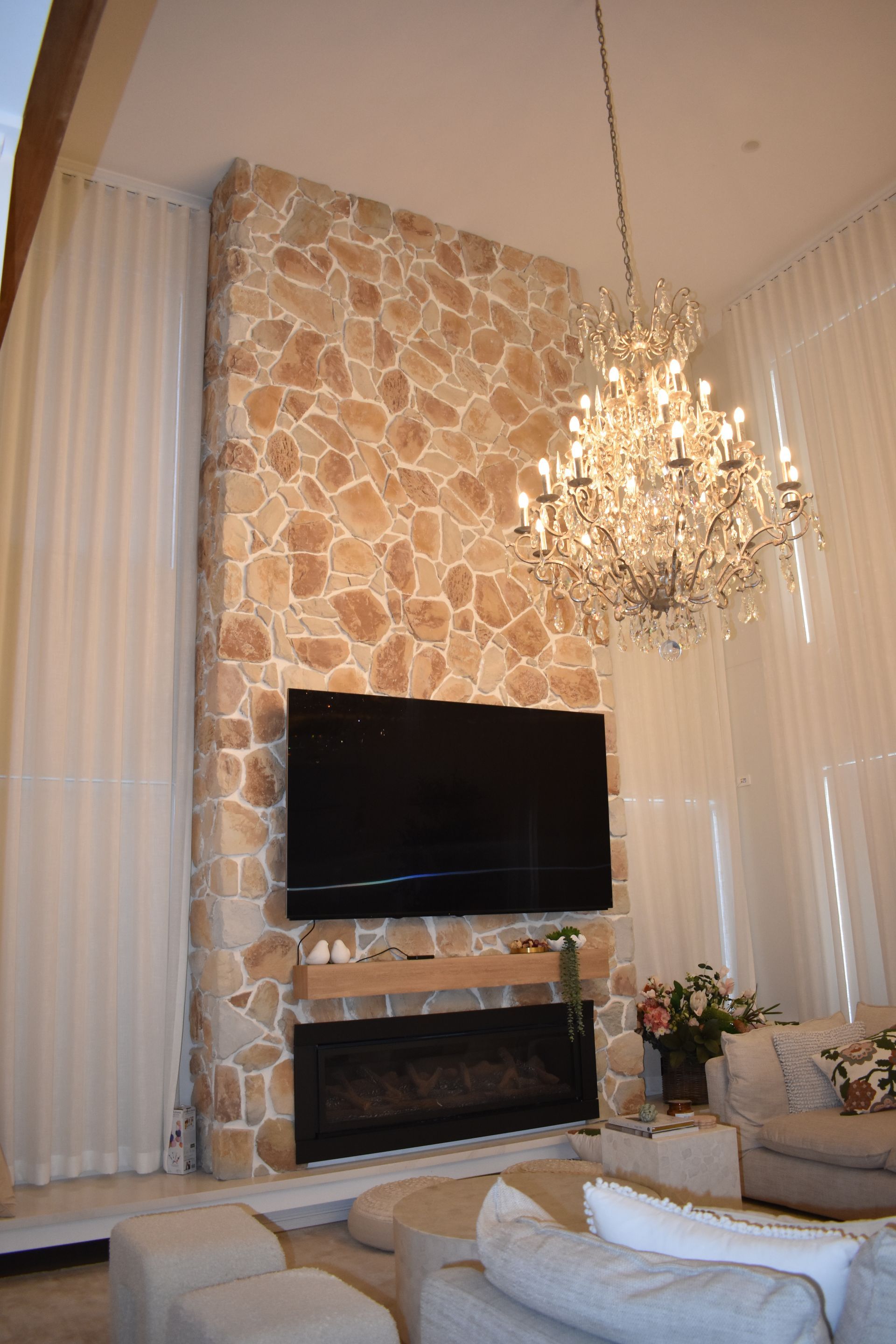 Elegant living room with stone fireplace, wall-mounted TV, chandelier, and tall white curtains