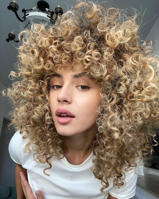A woman with curly blonde hair is wearing a white t-shirt.