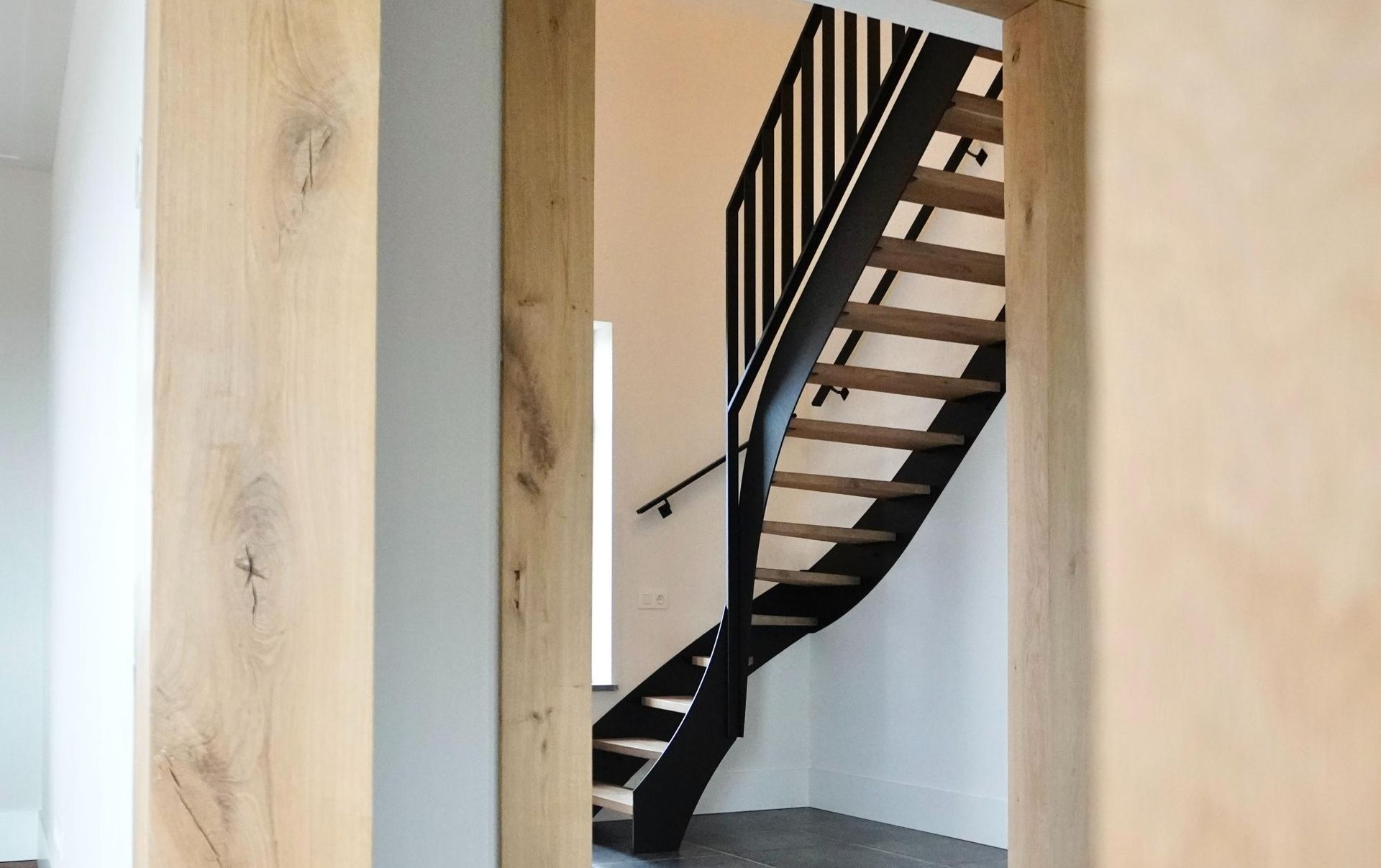 Houten trap met zwarte leuning en witte muren, zichtbaar vanuit een deuropening met houten afwerking.