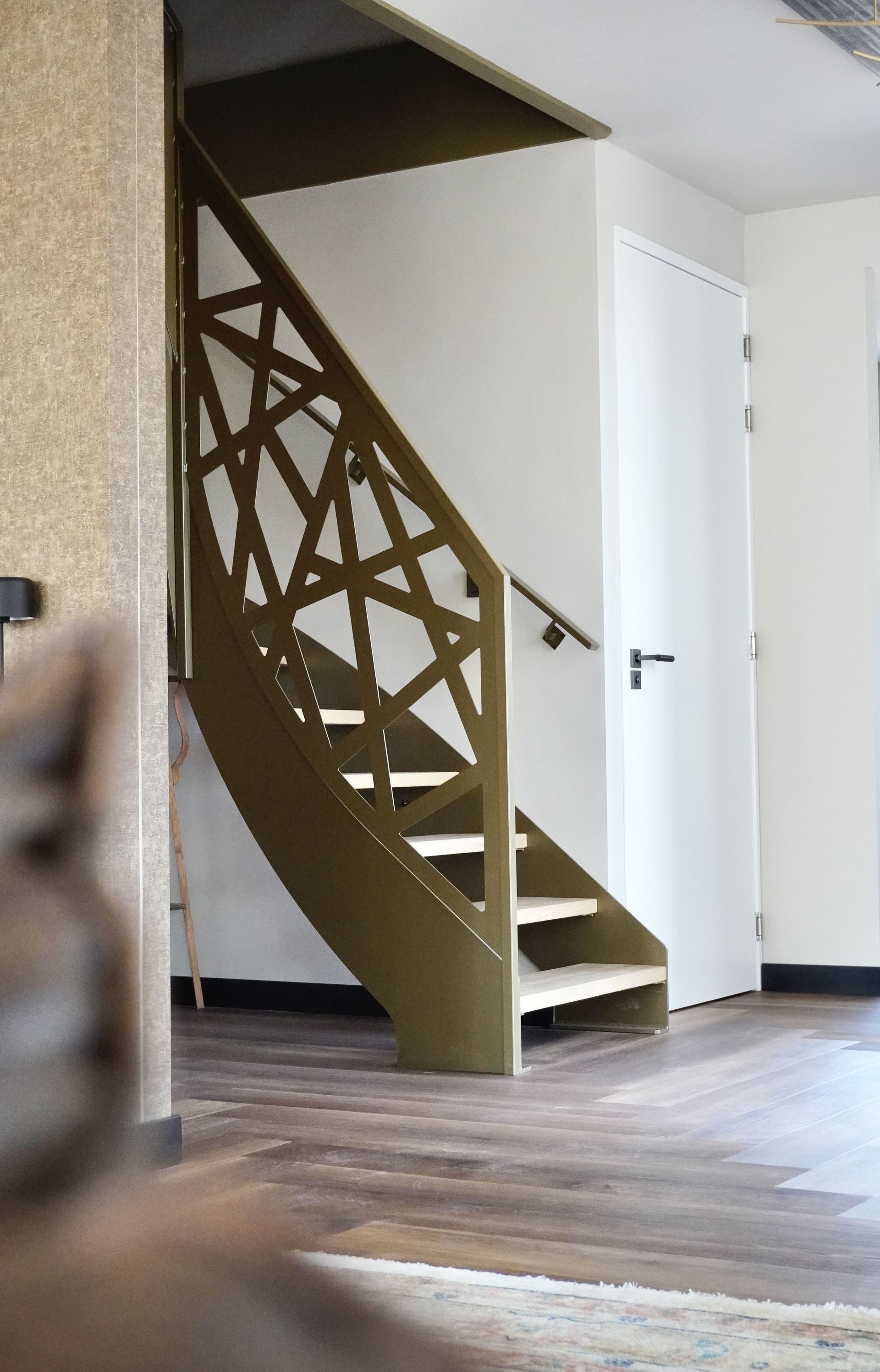 Een gebogen trap met een ingewikkeld geometrisch ontwerp, lichte houten treden en een witte deur op de achtergrond.