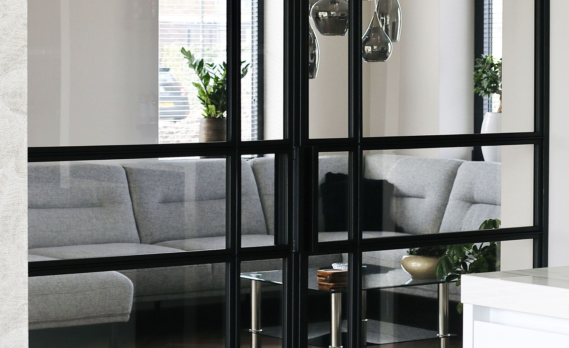 Zwarte glazen deuren scheiden een woonkamer met een grijze bank en decoratieve planten.