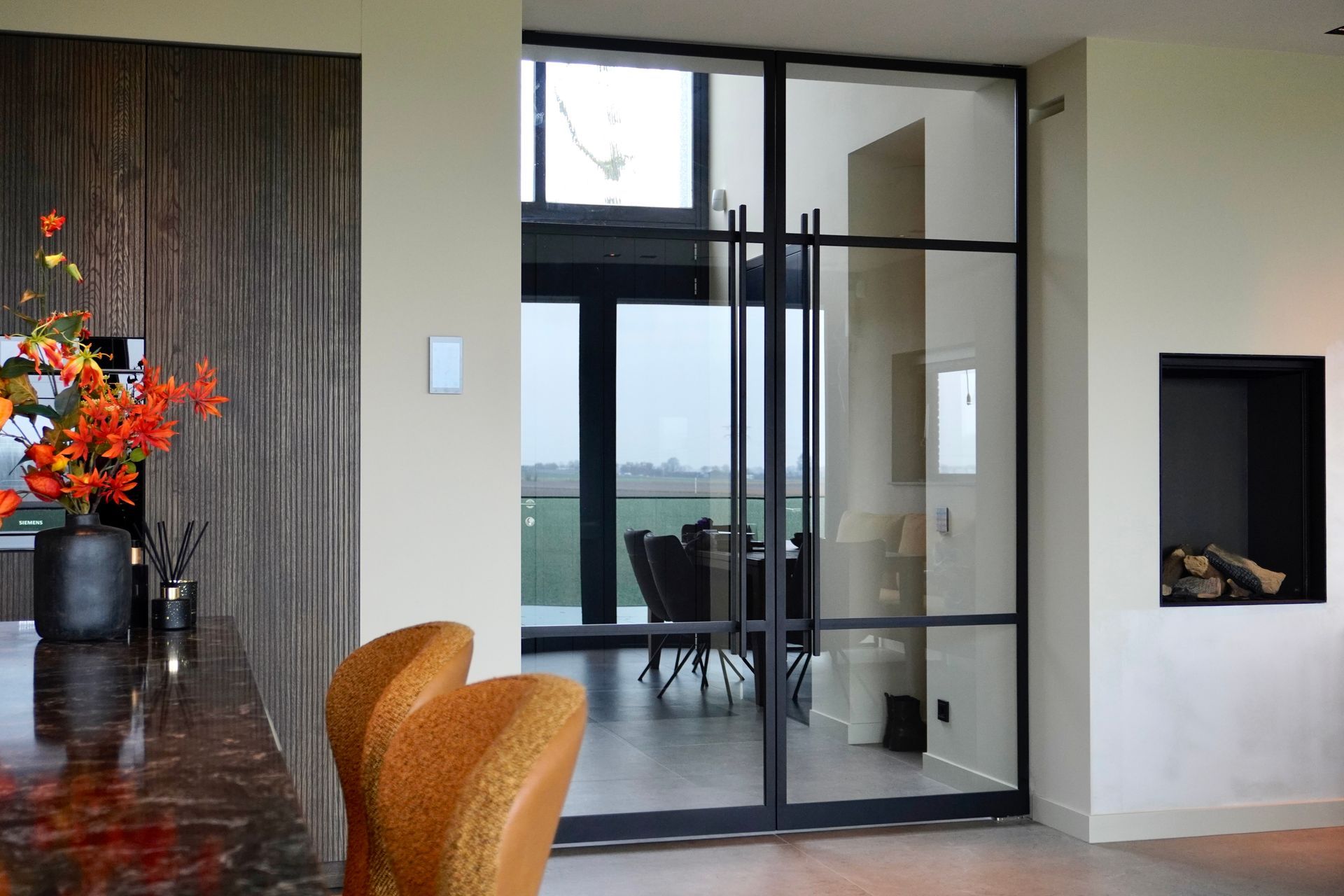 Modern interieur met glazen deuren, eethoek, open haard en oranje bloemstuk.