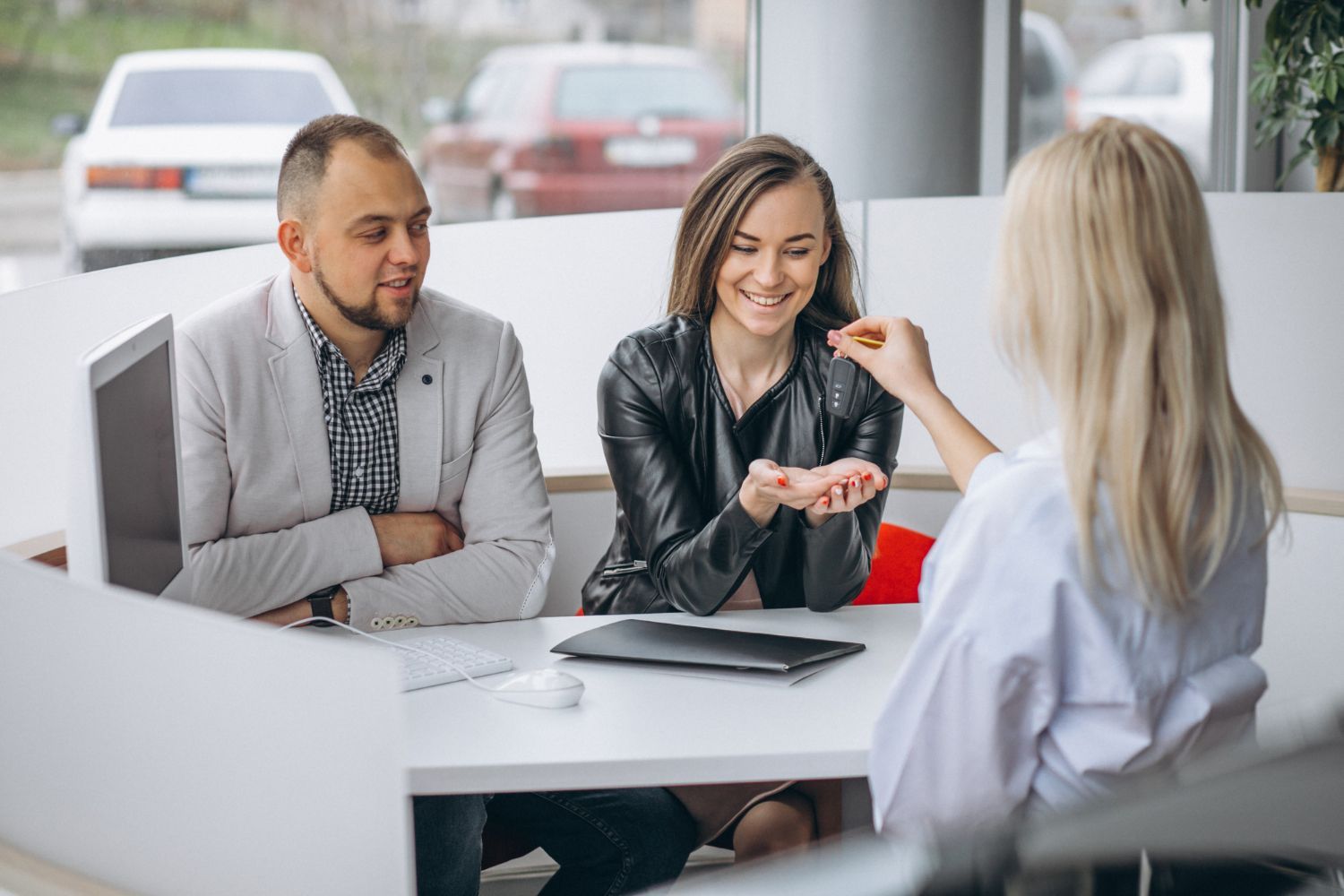 A woman receives keys from an employee at a desk inside a car dealership, with a man sitting next to her.