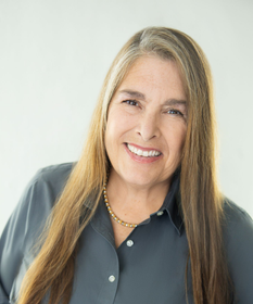 A person with short brown hair wearing a white button-down shirt and a pearl necklace, posing against a neutral backdrop.
