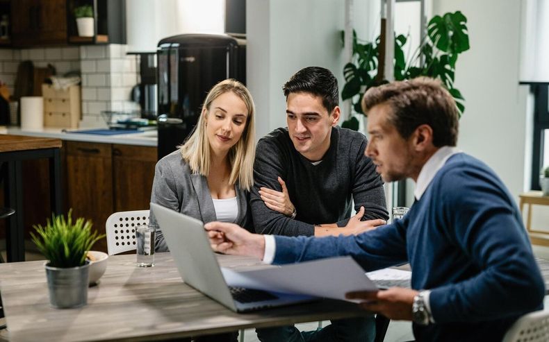 Three people sit at a table looking at a laptop and documents in a home kitchen setting.