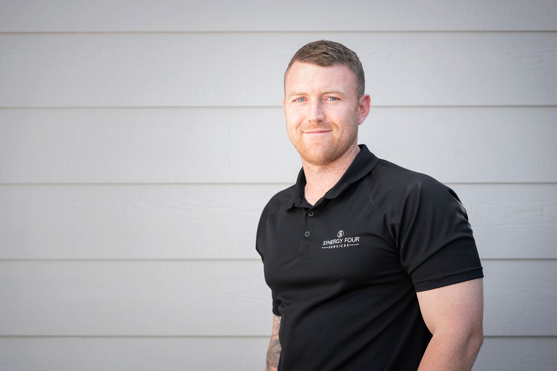 A man with a beard wearing a black polo shirt is standing in front of a white wall.