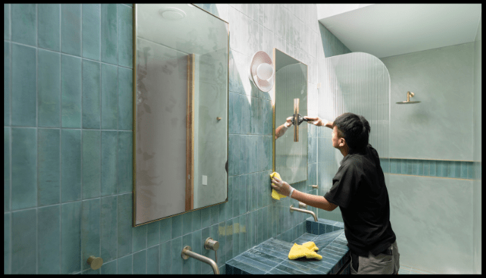 Person cleaning a mirror in a tiled bathroom using a spray bottle and cloth.