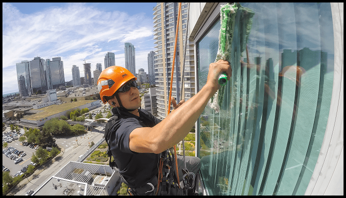 Professional window cleaner using rope access technique on a high-rise building with city skyline in the background.