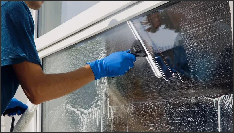 Worker wearing blue gloves using a squeegee to clean a large window with soapy water.