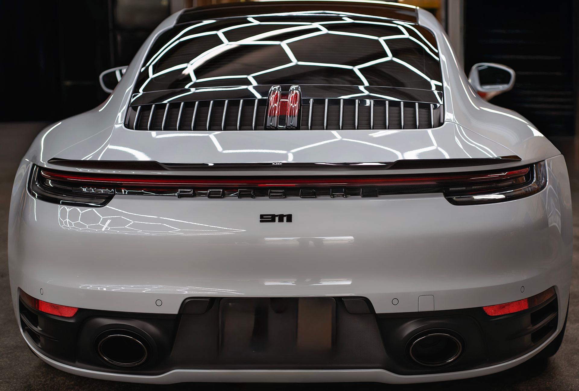 White Porsche 911 rear view, showcasing taillights and exhaust pipes. Reflective overhead lighting.