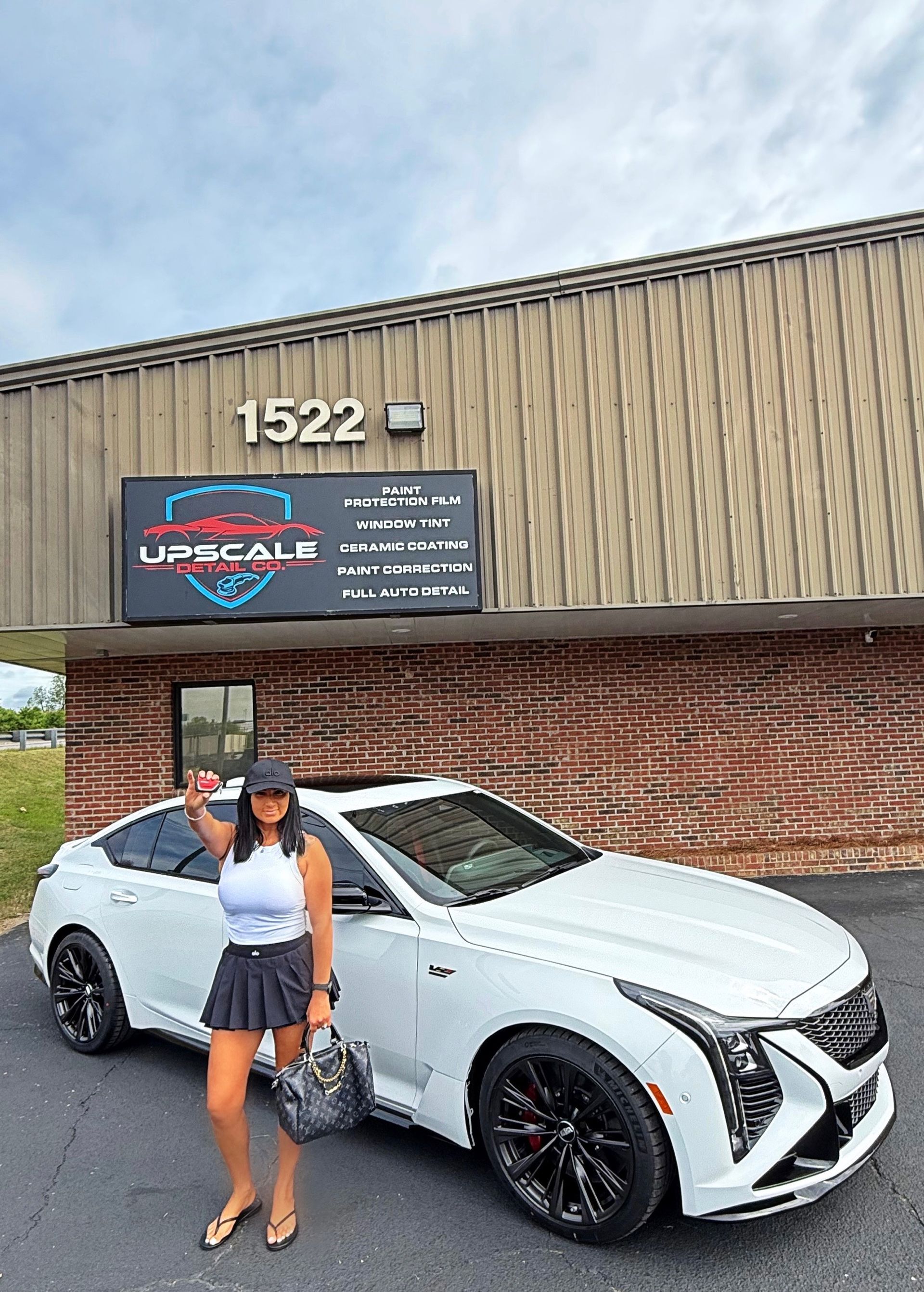 Woman posing beside a white sports car in front of a brick building with sign 1522.