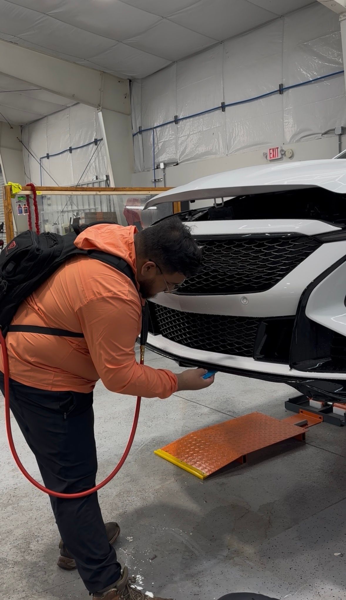 Person in an orange jacket spraying the rear of a white car in a workshop.