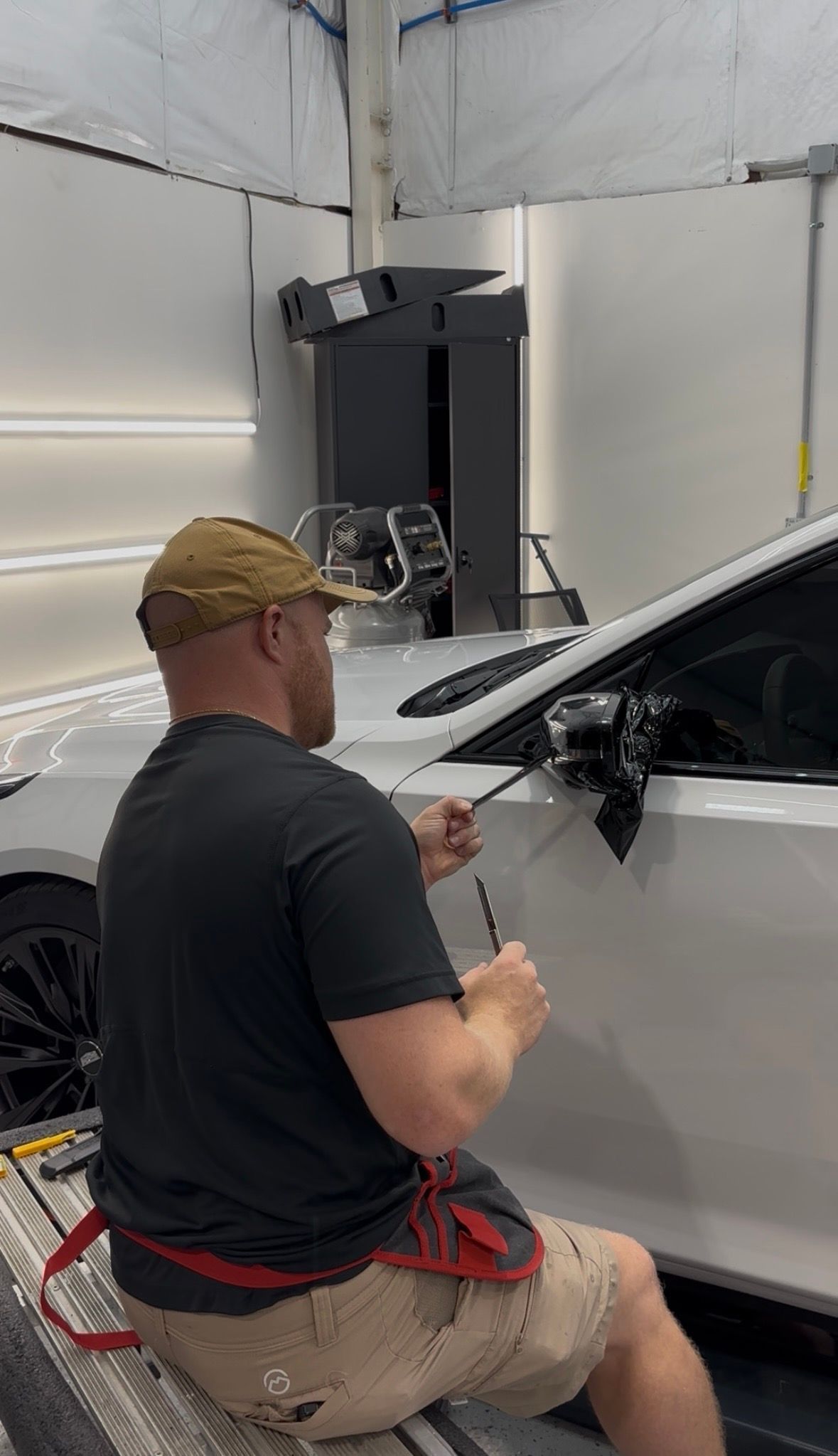 Mechanic working on a white SUV in a garage, using a tool near the side mirror.