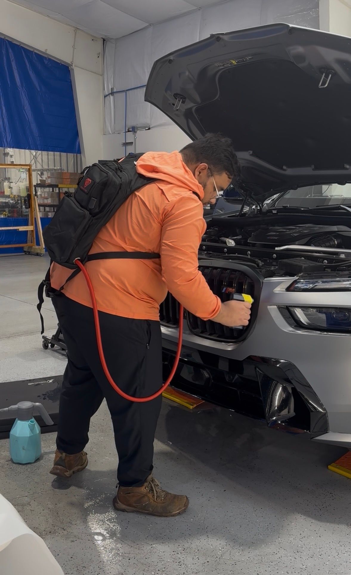 Mechanic in orange jacket inspecting the front of a car with its hood open in a garage