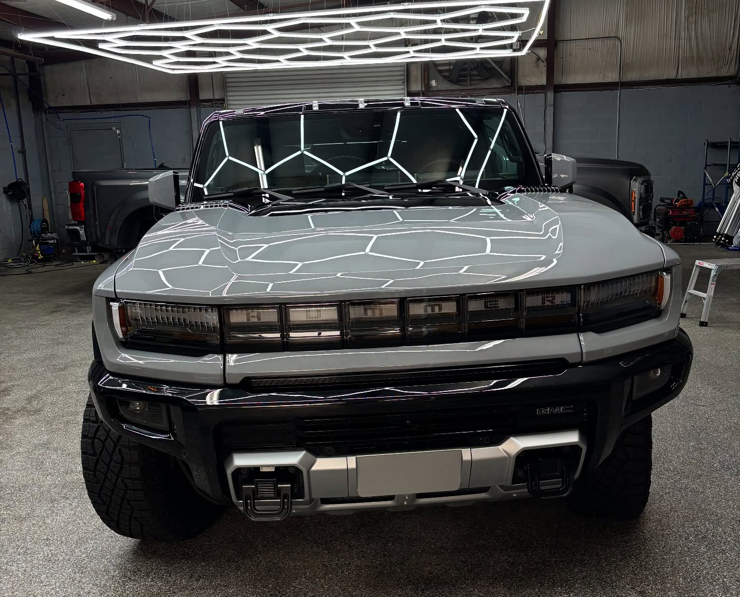 Gray GMC Hummer EV truck, front view, under hexagonal lights in a garage.