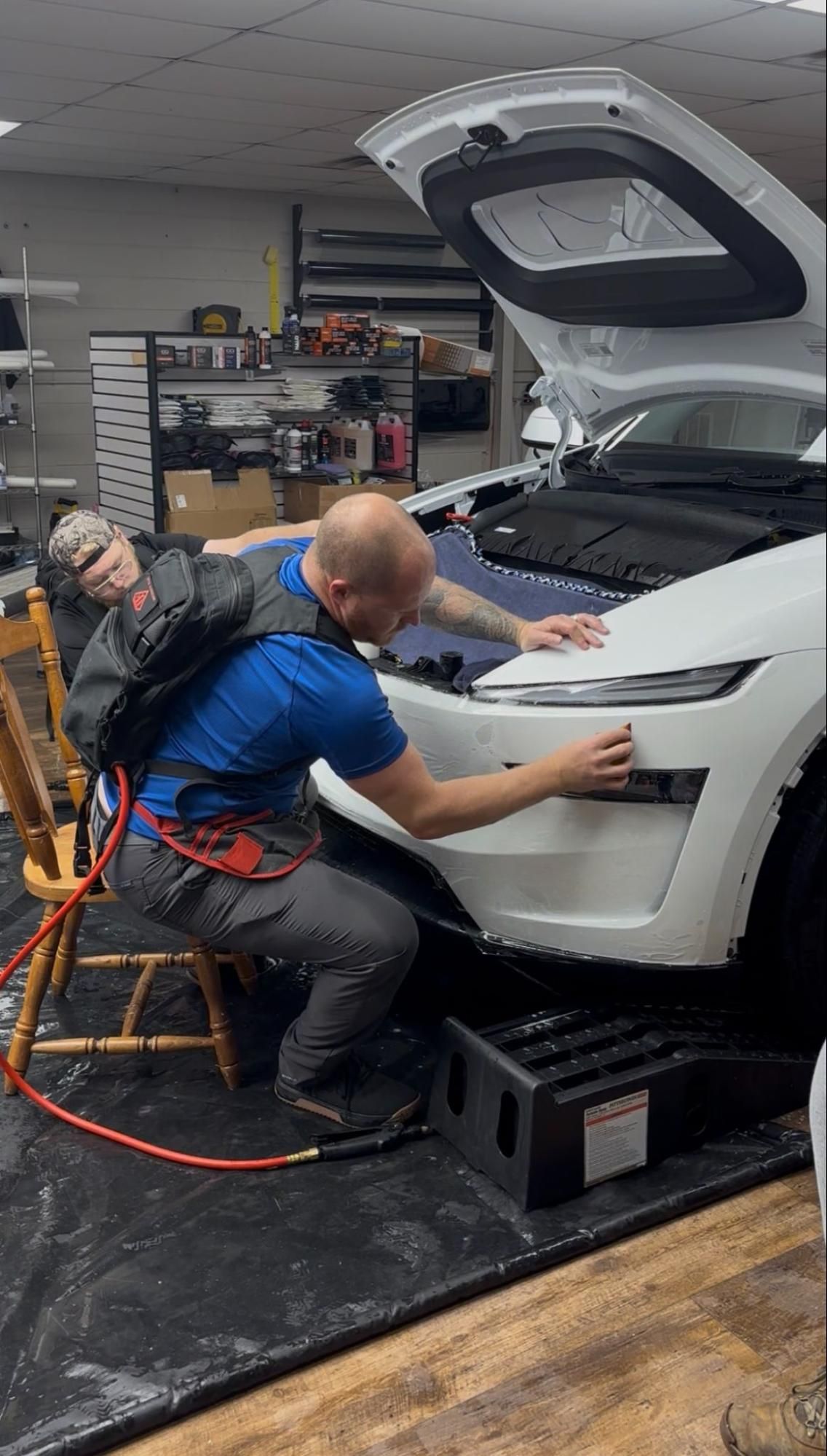 Mechanic inspecting a white car in a garage with the hood open, using tools near the front bumper
