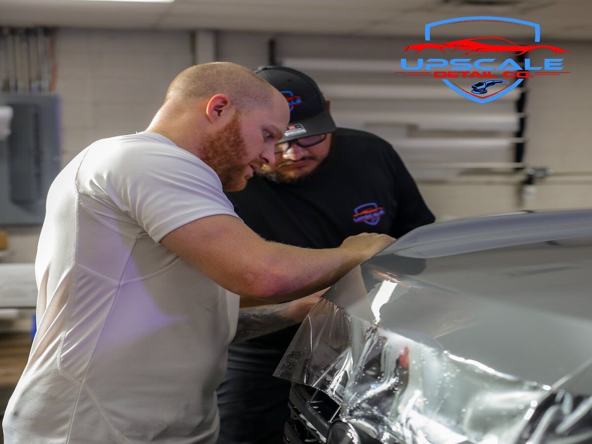 Person applying window tint to a car, using a heat gun and squeegee in a garage.